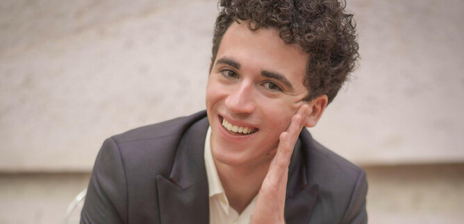 A young man with curly hair smiles relaxed. He is wearing a suit and has his hand on his face. | © privat