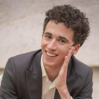 A young man with curly hair smiles relaxed. He is wearing a suit and has his hand on his face. | © privat