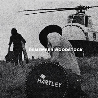 A group of people is moving in a field while a helicopter hovers in the background. In the center of the scene, there is a large suitcase with the inscription "HARTLEY." | © Rockhouse Salzburg