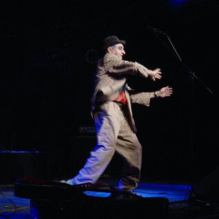 An artist on stage is performing. He is wearing a suit and a hat while colorful lights are shining. | © Roswitha Pross