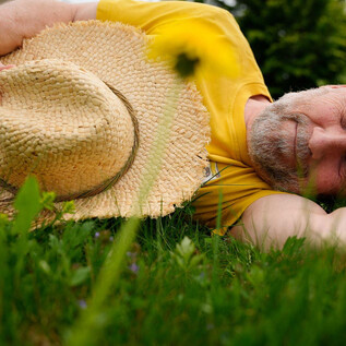 A man is lying relaxed on the grass wearing a sun hat. He enjoys nature in a quiet setting. | © Tom-Tom Lachmayr