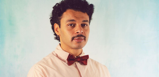 A man with curly hair is wearing a pink shirt and a burgundy bow tie. The background is bright and monochromatic. | © Isabella Aust
