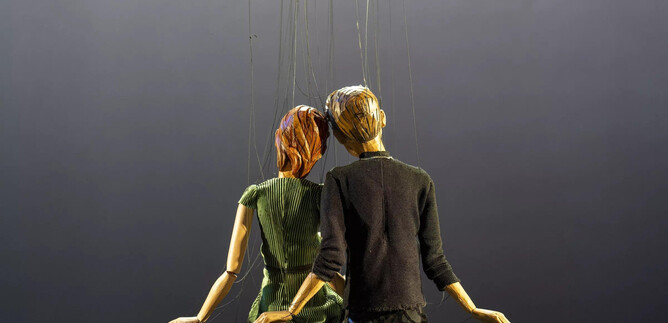 A figure or puppet of a couple sits together, held by strings. The background is dark and inconspicuous. | © Bernhard Müller