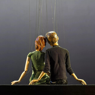 A figure or puppet of a couple sits together, held by strings. The background is dark and inconspicuous. | © Bernhard Müller