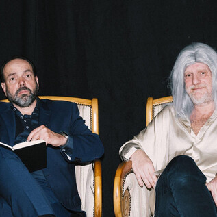 Two men are sitting in elegant chairs. The man on the left side is holding a notebook, while the other is wearing a long, gray wig. | © Ingo Pertramer