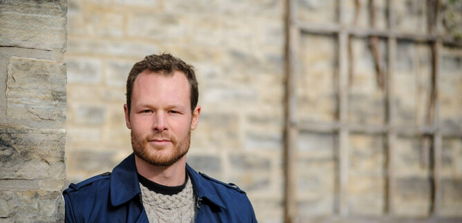 A man with blonde hair stands by a stone wall. He is wearing a blue coat and looking directly at the camera. | © Bill Mccarthy