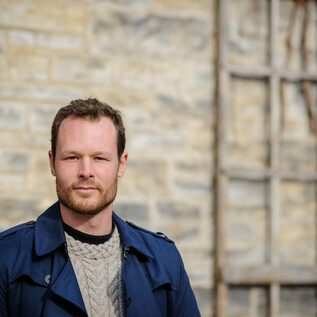 A man with blonde hair stands by a stone wall. He is wearing a blue coat and looking directly at the camera. | © Bill Mccarthy