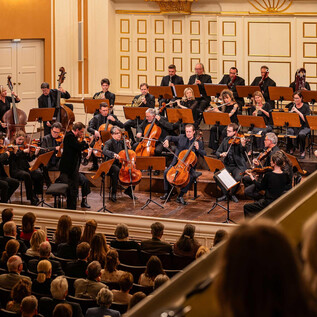 An orchestra is playing in an elegant hall in front of an enthusiastic audience. The musicians are gathered on stage and are focusing on the conductor. | © Ebihara Photography