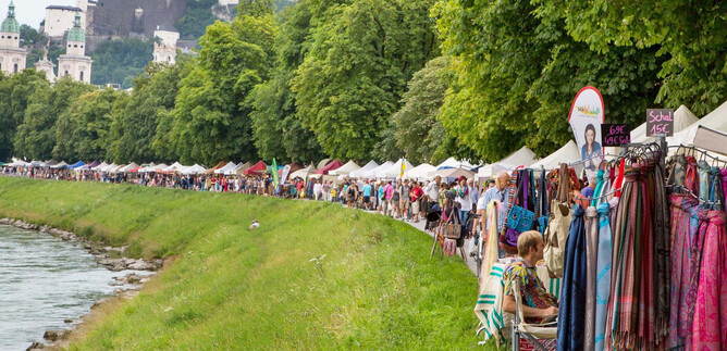 A market with colorful stalls along a river. In the background, there are green trees and a city view with historic buildings.