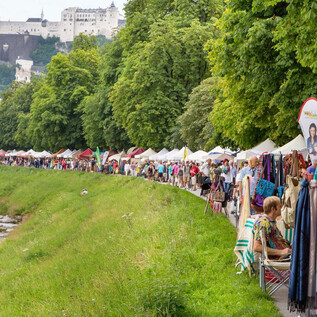 A market with colorful stalls along a river. In the background, there are green trees and a city view with historic buildings.