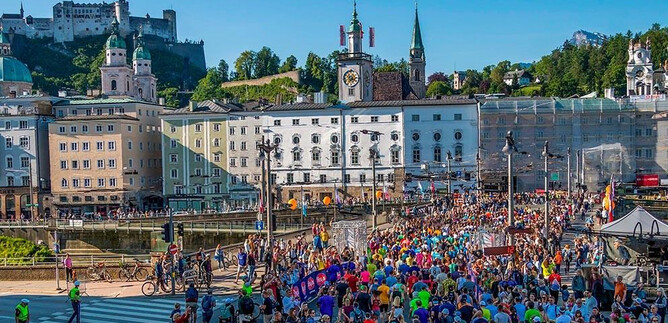 A lively event with many people in colorful T-shirts on a busy street. In the background, historic buildings and a castle can be seen. | © Salzburg Marathon_Christian Köhler