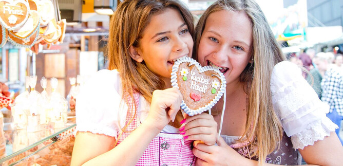 Two girls smile and hold a heart made of gingerbread while wearing traditional costumes. In the background, more festivities can be seen. | © wildbild