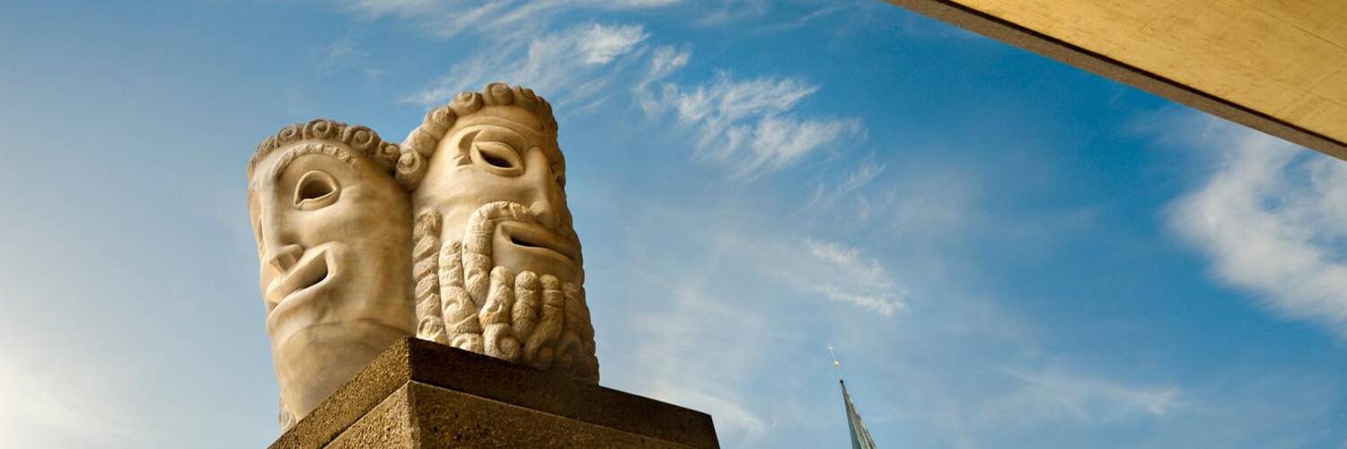 Two artistic stone heads stand on a pedestal. In the background, a bright sky with a few clouds and a church tower can be seen. | © Luigi Caputo