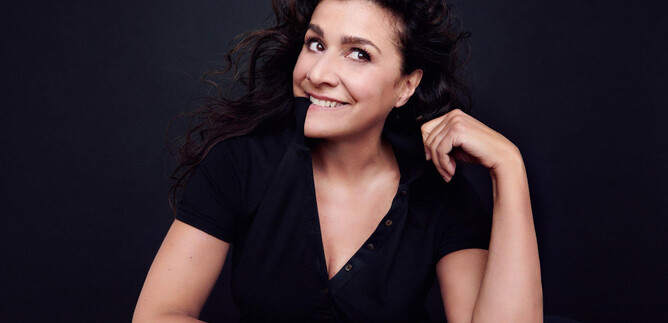 A smiling woman with curly hair sits in front of a dark background. She is wearing a black top and looks friendly. | © Kristian_Schuller_Decca