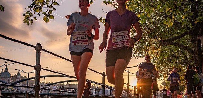 Two women jog in the twilight along a path surrounded by trees. In the background, a bustling scene with more runners and a beautiful cityscape is visible. | © Salzburger Frauenlauf_Alexander Schwarz