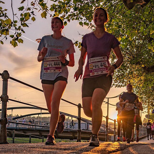 Two women jog in the twilight along a path surrounded by trees. In the background, a bustling scene with more runners and a beautiful cityscape is visible. | © Salzburger Frauenlauf_Alexander Schwarz