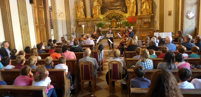 A concert hall with many spectators listening to an ensemble of four musicians. The decoration is magnificent, with golden statues and a festive ambiance.