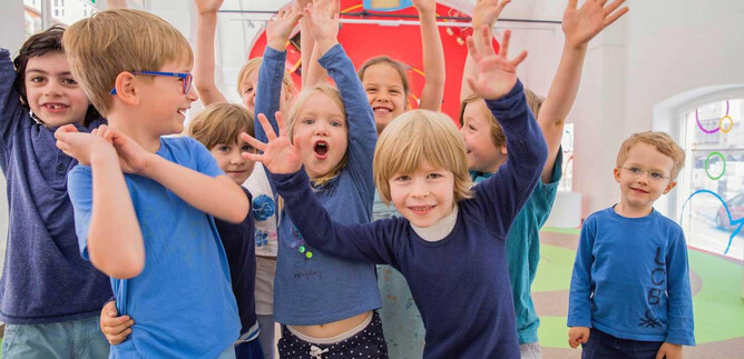 A group of happy children stands together and raises their hands in the air. They are wearing casual clothes in various shades of blue and are laughing.