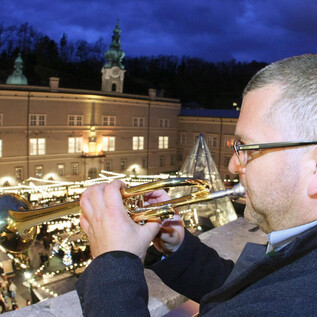 A man is playing trumpet on a balcony. In the background, festively illuminated stalls and crowds of people can be seen. | © www.christkindlmarkt.co.at, Salzburg
