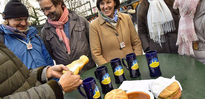 A cheerful group of people is standing around a table with food and drinks. On the table, there are rolls and a plate with warm soup. | © salzburg-experience.at