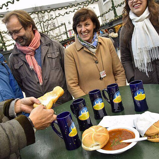 A cheerful group of people is standing around a table with food and drinks. On the table, there are rolls and a plate with warm soup. | © salzburg-experience.at