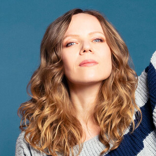A person with curly hair is wearing a striped sweater. They are confidently looking into the camera and raising one arm. | © Anne Wilk