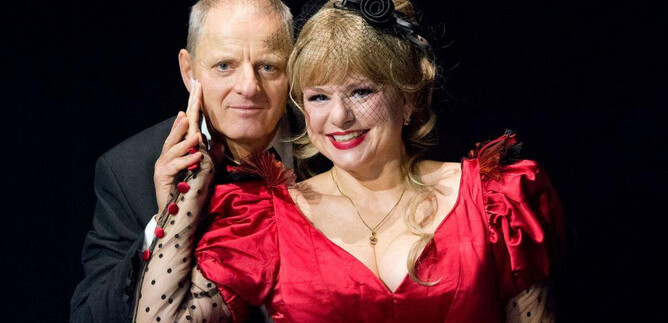 A man and a woman are posing together on a stage. The woman is wearing a red dress and has a cheerful demeanor. | © Christian Treweller