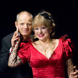 A man and a woman are posing together on a stage. The woman is wearing a red dress and has a cheerful demeanor. | © Christian Treweller