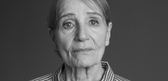 An older woman with a contemplative expression stands in front of a neutral background. She is wearing a striped shirt and has a gentle gaze. | © Armin Smailovic