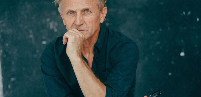 A thoughtful man with blonde hair is sitting with one hand on his chin. The background is dark and simple. | © Nowy Teatr
