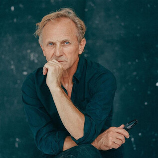 A thoughtful man with blonde hair is sitting with one hand on his chin. The background is dark and simple. | © Nowy Teatr