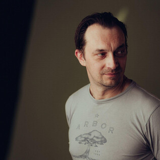 A man stands slightly sideways and looks thoughtful. He is wearing a gray T-shirt with a print. | © Joel Heyd