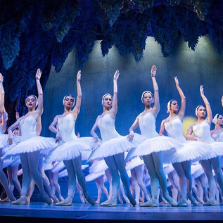 A group of dancers in white ballet costumes. They are performing a synchronized dance on a stage with a dramatic background. | © International Festival Ballet