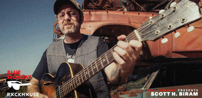 A man is playing guitar in a rustic setting with old cars in the background. He is wearing a cap and has a relaxed expression on his face. | © Rockhouse Salzburg