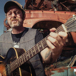A man is playing guitar in a rustic setting with old cars in the background. He is wearing a cap and has a relaxed expression on his face. | © Rockhouse Salzburg