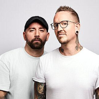 Two men are standing next to each other in front of a bright background. Both are wearing casual clothing and have tattoos. | © Pascal Riesinger