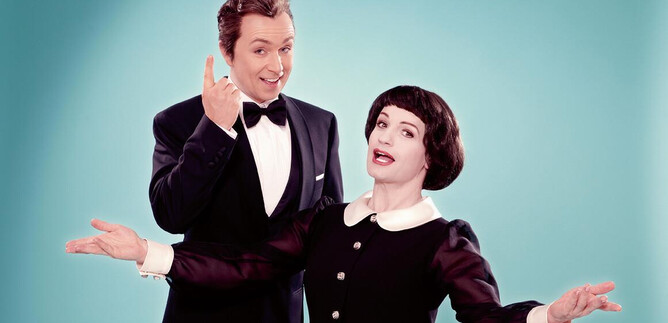A man in a tuxedo and a woman in a black dress are smiling in front of a light blue background. They seem to be announcing an entertaining performance. | © FOKKE