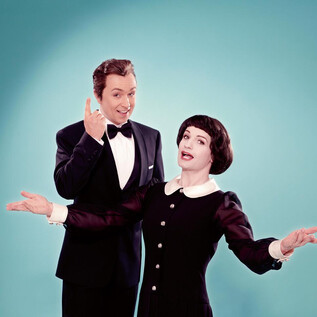 A man in a tuxedo and a woman in a black dress are smiling in front of a light blue background. They seem to be announcing an entertaining performance. | © FOKKE