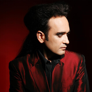 A man with dark, voluminous hair is wearing a red suit. The background is in a vibrant red. | © Rockhouse Salzburg