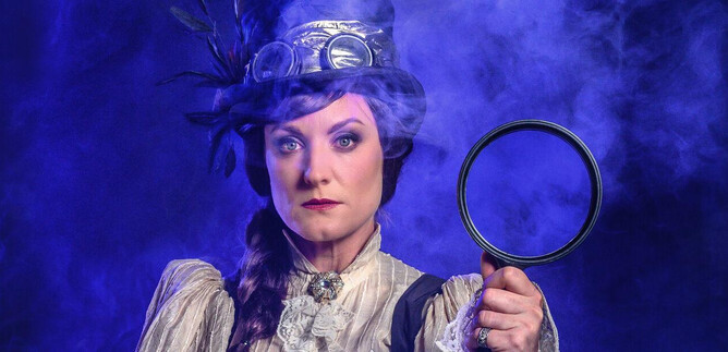 A woman in historic clothing holds a magnifying glass in her hand. The background is mystical with blue smoke. | © SLT / Tobias Witzgall
