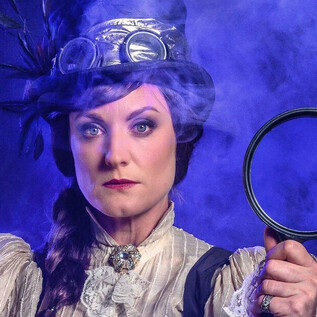 A woman in historic clothing holds a magnifying glass in her hand. The background is mystical with blue smoke. | © SLT / Tobias Witzgall