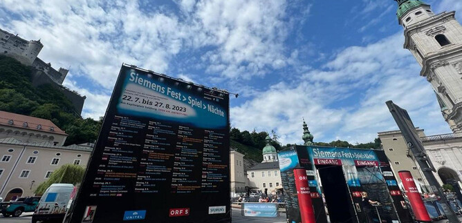 An event venue with large information walls for the Siemens festival takes place outdoors. In the background, historical buildings and a beautiful sky scene can be seen.