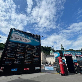 An event venue with large information walls for the Siemens festival takes place outdoors. In the background, historical buildings and a beautiful sky scene can be seen.