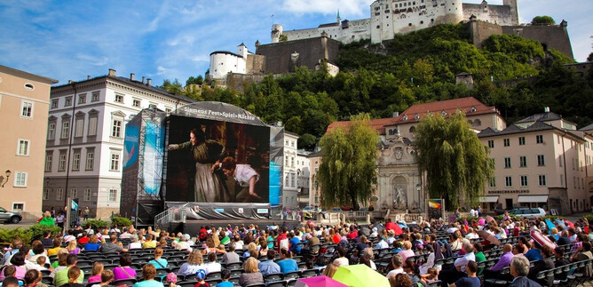 A large audience is sitting on chairs in a city while watching a performance on a screen. In the background, an impressive castle can be seen on a hill. | © Siemens AG Österreich