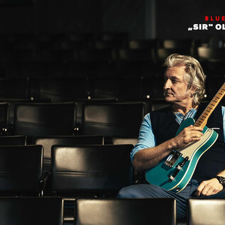 A male musician is sitting on an empty row of benches holding an electric guitar in his hand. He is wearing a blue shirt and has gray hair. | © Bernd Grosseck