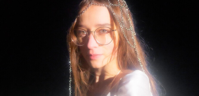 A young woman with glasses and a sparkling headpiece looks directly at the camera. The background is black, which emphasizes her expression. | © Michelle Rassnitzer