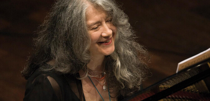 A smiling woman plays the piano. Her long, gray hair falls in curls over her shoulders. | © Adriano Heitman