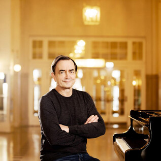 A musician sits smiling in front of a piano. In the background are elegant rooms with warm light. | © Julia Wesely