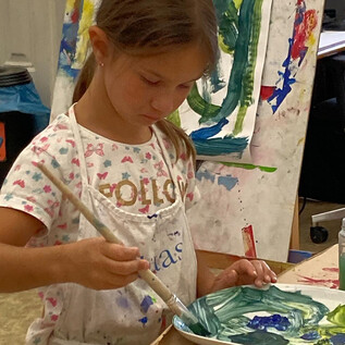 Une petite fille peint soigneusement avec un pinceau sur une palette. En arrière-plan, des peintures colorées sont visibles. | © DomQuartier