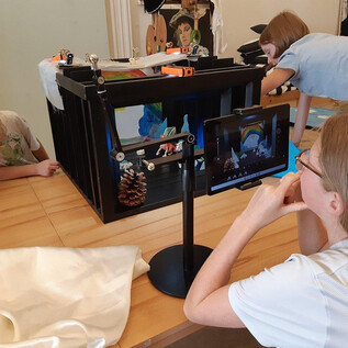 Children experiment with a creative film set environment. An adult observes them as they work with props and a camera. | © DomQuartier